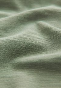 Švelni, tekstūruota medžiaga pastelinių žalių tonų, su subtiliu mezgimo raštu ir švelniais klostėmis, rodančiomis lankstumą ir krintančių savybių.
