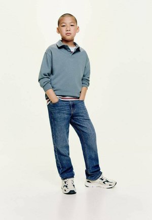 Polo shirt azzurro chiaro a maniche lunghe con colletto, abbinato a jeans blu e sneaker bianche con dettagli neri. Sfondo semplice.