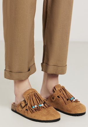 Scarpe slip-on in suede color tan con design frangiato, decorate con perline colorate e una fibbia laterale. Suola in gomma nera, abbinate a pantaloni cropped beige.