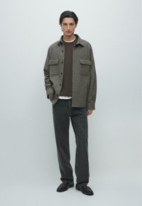 Grå skjortejakke i uldblanding med to forlommer, en brun tekstureret sweater under og mørke jeans, komplet med sorte loafers.