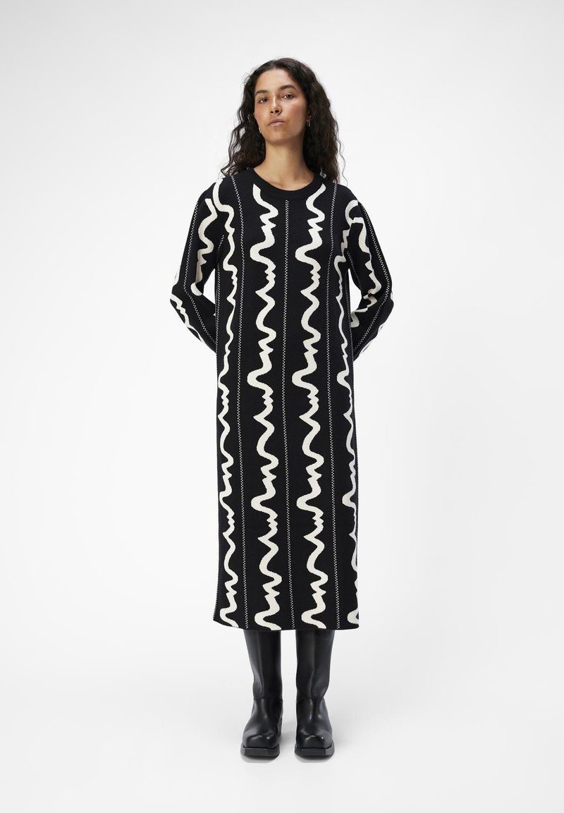 Robe longue noire avec des motifs ondulés blancs. Dispose d'un col rond et de manches longues. Assortie avec des bottes noires jusqu'aux genoux.