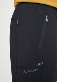 Pantalons techniques noirs avec poches latérales zippées, dotés d'une finition mate, d'éléments de design angulaires et d'un logo situé sur le bas de la jambe.