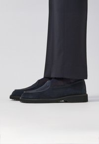 Dark navy slip-on shoes with smooth suede texture, rounded toe, and subtle stitching detailing. Worn with tailored dark pants.