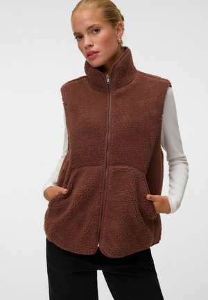 Femme portant un gilet polaire sans manches marron zippé avec poches, par-dessus un haut blanc à manches longues, sur un fond uni.