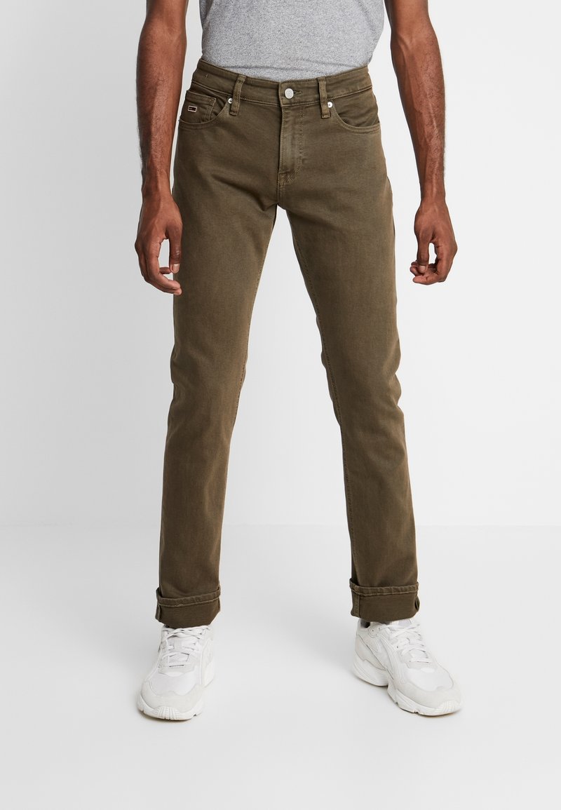 Homme portant un jean vert olive avec des revers et des baskets blanches, debout devant un fond gris clair uni.