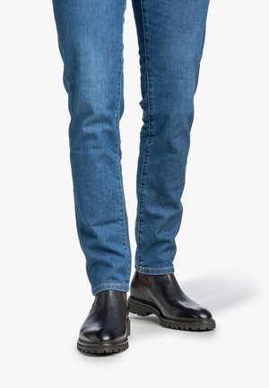 Une personne portant un jean en denim bleu et des bottines Chelsea en cuir noir se tenant sur un fond blanc.