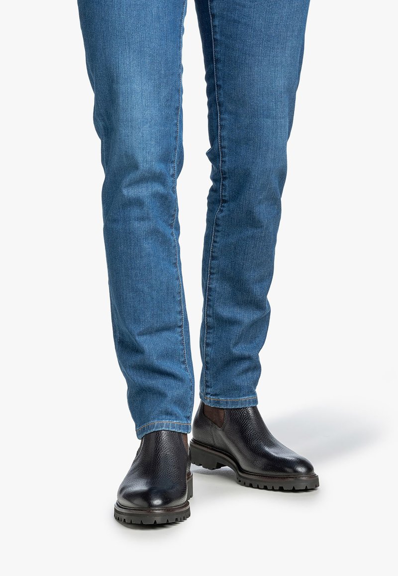 Persona vestida con jeans de mezclilla azul y botas Chelsea de cuero negras de pie sobre un fondo blanco.