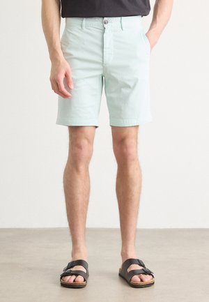 Mand iført lyseblå shorts, sorte sandaler med spænder og en mørk skjorte, står på en glat gulv mod en ensfarvet væg.