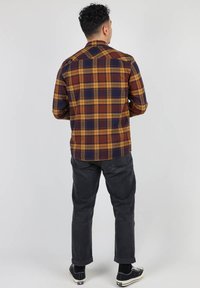 Chemise à carreaux marron, jaune et marine, fabriquée en tissu tissé ; présente un col pointu et un détail de empiècement arrière unique.