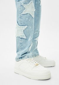 Jeans de mezclilla azul claro con parches de estrellas deshilachados y manchas de pintura blanca, llevados con zapatillas blancas con cordones sobre un fondo blanco liso.