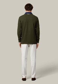 Zielony sweter z dzianiny z długimi rękawami, granatowym kołnierzem i łatami na łokciach; w połączeniu z białymi spodniami i brązowym obuwiem. Gładkie tło.