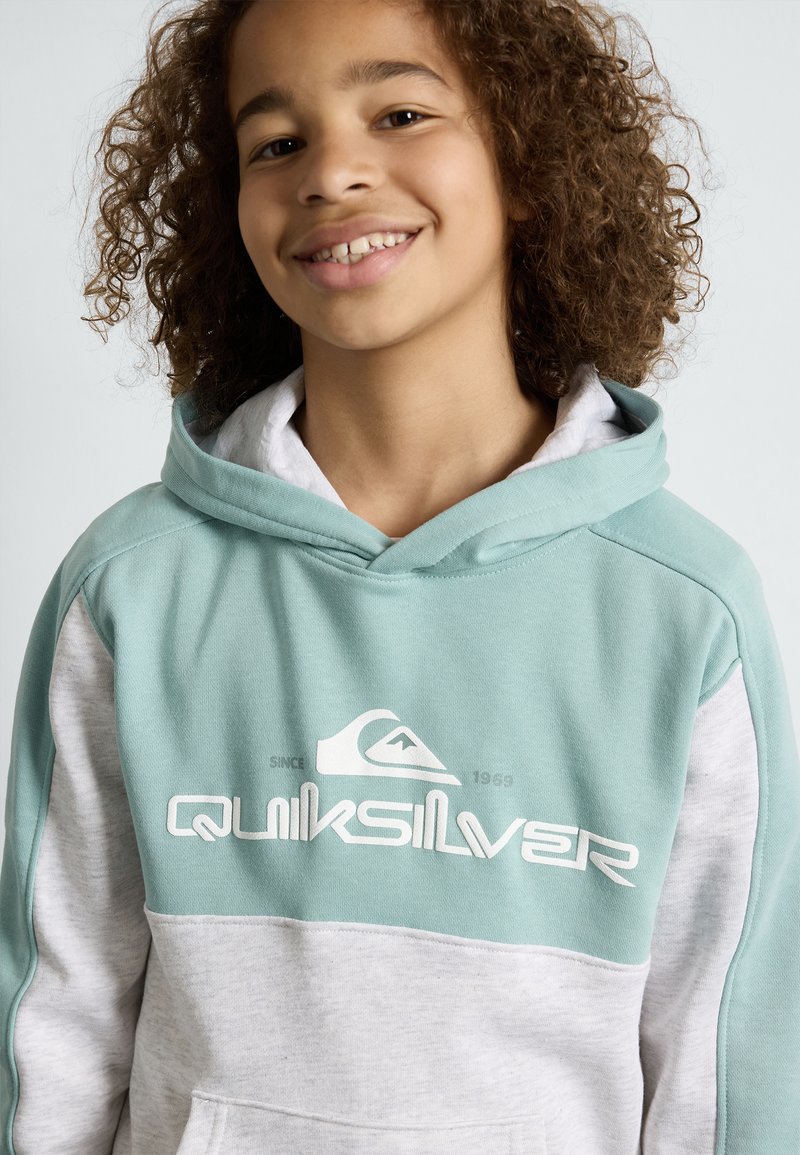 Kardigan s kapuljačom u tirkiznom gornjem dijelu i sivom donjem dijelu. Sadrži Quiksilver logo u bijeloj boji, mekani materijal i ležeran kroj.