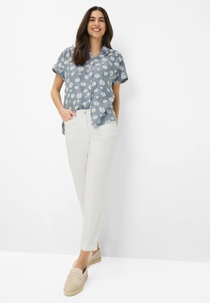 Woman smiling, wearing short-sleeve blue floral button-up shirt, white pants, and beige slip-on shoes, standing against white background.