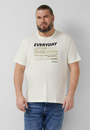 Mężczyzna z brodą w białej koszulce z napisem „EVERYDAY” i niebieskich dżinsach, stojący na jednolitym jasnoszarym tle.