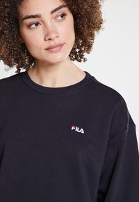 Felpa nera con scollatura rotonda, polsini a coste e un piccolo logo ricamato bianco e rosso sul lato sinistro del petto.