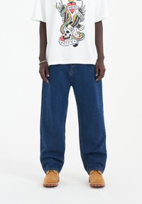 Bílý tričko s barevným grafickým designem obsahující lebku, hada a orla. Spárováno s volnými modrými džínami a žlutými botami.