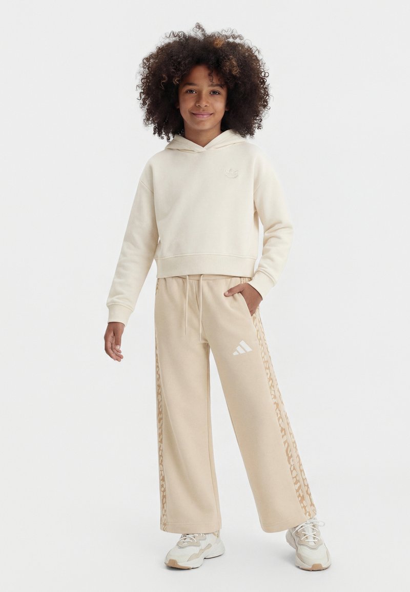 Παιδί με σγουρά μαλλιά, φορώντας μπεζ φούτερ με κουκούλα, παντελόνια με φαρδιά πατζάκια και πλευρικά σχέδια, και λευκά αθλητικά παπούτσια, στέκεται με το ένα χέρι στην τσέπη.