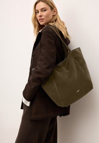 Olíva zöld vászontáska sima felülettel és két erős füllel. Diszkrét márkajelzés a fronton, semleges háttérrel.