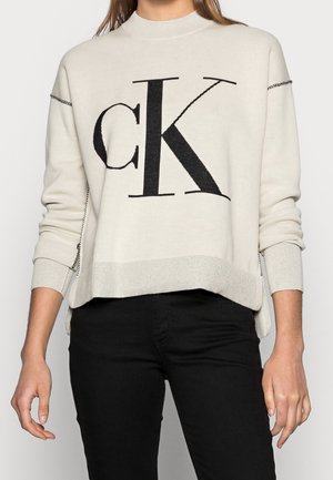 Personne portant un pull crème avec le logo noir "cK" sur le devant et un pantalon noir, debout devant un fond clair uni.