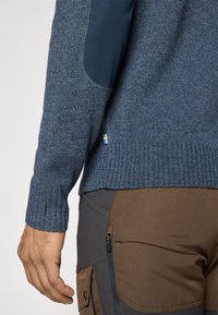 Prim-plan al unei persoane care poartă un pulover tricotat albastru cu petice la coate și pantaloni de exterior maro, care au un buzunar cu fermoar și un buzunar cu clapetă.