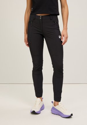 Black stretchy pants with a slim fit, featuring zippered pockets, subtle branding, and a smooth texture. Worn with light-colored sneakers.