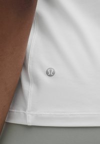 Lähedalt vaade inimese käsivarrele valgele sportlikule särgile, mille allääres on väike hall logo ja nähtavad õmbluse detailid.