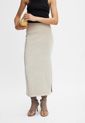 Beige stickad maxikjol med en sidoklyvning, tillsammans med en svart ärmlös topp och bruna högklackade skor med remmar, som håller en gul kuvertväska.