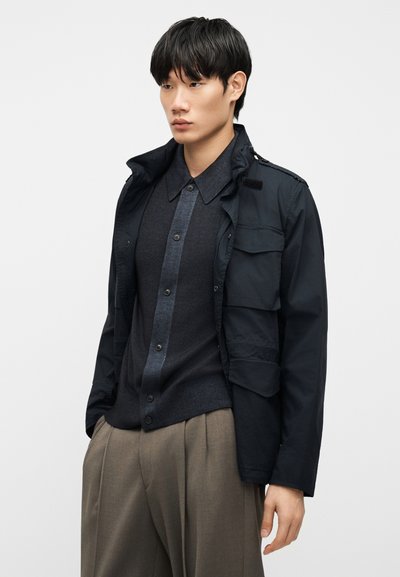 Jeune homme portant un cardigan sombre boutonné, une veste noire avec des poches avant, et un pantalon beige ample sur un fond uni.