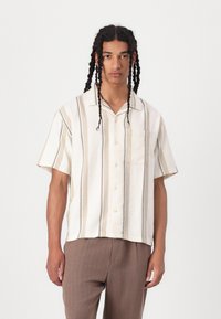 Chemise à manches courtes de couleur crème avec de fines rayures verticales en tan et bleu marine. Comprend un devant à boutons et une poche poitrine.