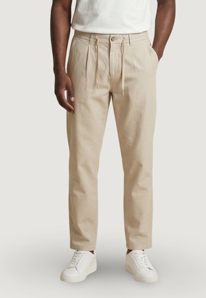 Uomo con pantaloni beige plissettati con vita a coulisse, sneakers bianche e t-shirt bianca, in piedi davanti a uno sfondo chiaro e uniforme.