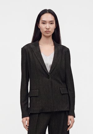 Femme aux longs cheveux noirs portant un blazer à motif chevrons foncé et un pantalon assorti sur un haut gris clair sur un fond uni.