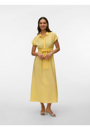 Kvinde stående iført en gul midikjole med korte ærmer, taljebælte og åbne højhælede sandaler.