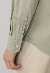 Camicia verde a maniche lunghe con polsini chiusi da bottoni, caratterizzata da pieghe sulle maniche e bottoni simili a perle. Tessuto liscio con finitura opaca.