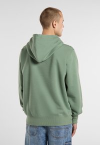 En man med kort hår bär en grön hoodie och ljusblå jeans, vänd bort från kameran mot en ljus bakgrund.