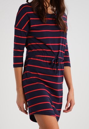 Nainen, joka on pukeutunut tummansiniseen mekkoon, jossa on punaiset vaakaraidat, 3/4-hihat ja vyötäröllä kiristysnyöri, seisoo yksiväristä taustaa vasten.