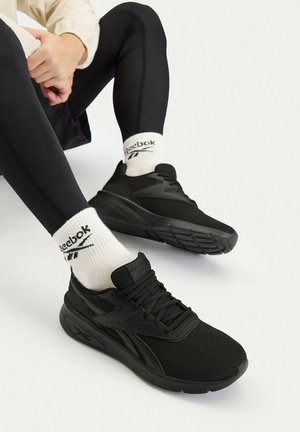 Person trägt schwarze Reebok-Sneaker, weiße Reebok-Socken und schwarze Leggings, sitzt mit zusammengelegten Händen in der Nähe der Knie vor weißem Hintergrund.