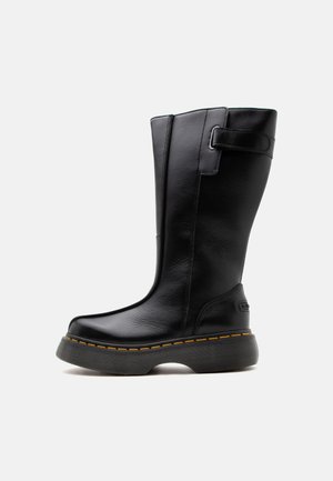 Botas altas de cuero negro con suela gruesa y costura amarilla, con una correa y hebilla cerca de la parte superior sobre un fondo liso y claro.