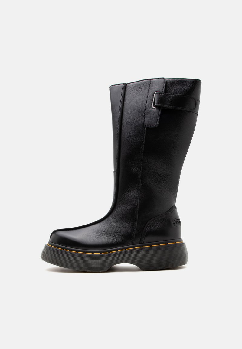 Botte haute en cuir noir avec semelle épaisse et coutures jaunes, dotée d'une sangle et d'une boucle près du haut, sur un fond clair uni.