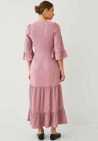 Lang roze jurk met driekwart uitlopende mouwen, textuurstof, een aansluitend lijfje en een gelaagde rok. Zwarte flats maken de look compleet.