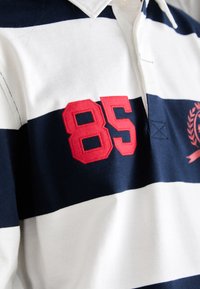 Polo-skjorte med marineblå og hvite horisontale striper, med et rødt brodert "85" på brystet og et klassisk kragedesign.