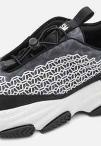 Černá a šedá atletická bota s texturovaným svrškem, bílým šestiúhelníkovým vzorem a elastickými tkaničkami. Má polstrovanou bílou podrážku a gumový grip.