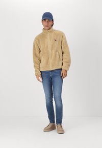 Chaqueta de forro polar beige con cuello alto y cremallera completa, gorra azul, jeans azules y zapatos beige. La chaqueta cuenta con un pequeño logo en el pecho.