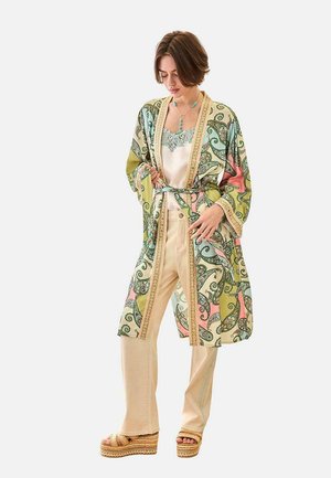 Mujer con un kimono estampado en verde, rosa y beige con cinturón, sobre pantalones crema, blusa de seda y sandalias de plataforma, posando frente a un fondo blanco.
