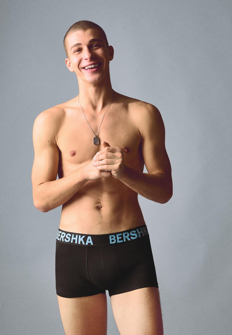 Jeune homme souriant, torse nu, portant un boxer noir avec une ceinture visible marquée "BERSHKA", debout devant un fond gris uni.