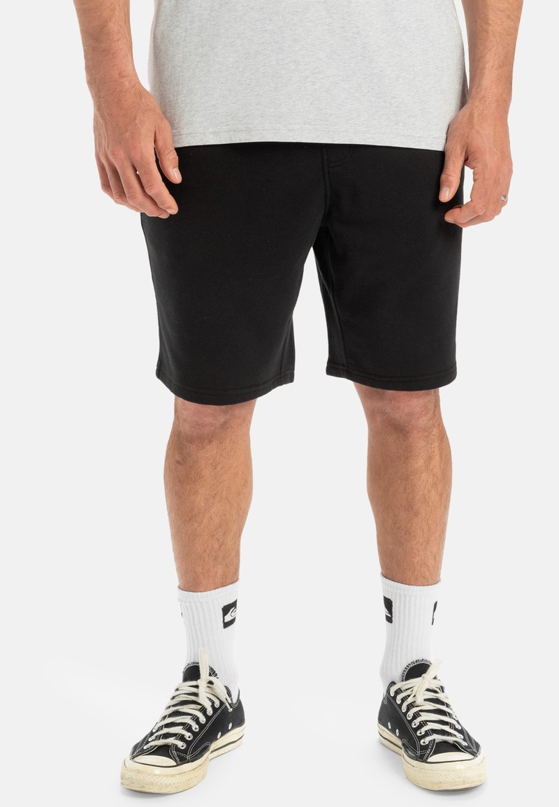 Pantalones cortos de algodón negros con un corte relajado, que terminan justo por encima de la rodilla, combinados con calcetines blancos hasta la altura del tobillo y zapatillas negras.