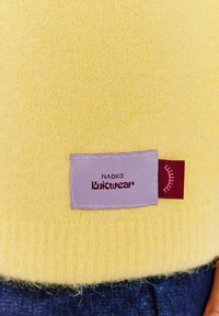 Žlutý pletený svetr s jemnou texturou, na němž je fialová a vínová etiketa s nápisem "Naoko Knitwear". Pozadí je tmavě modrý denim.