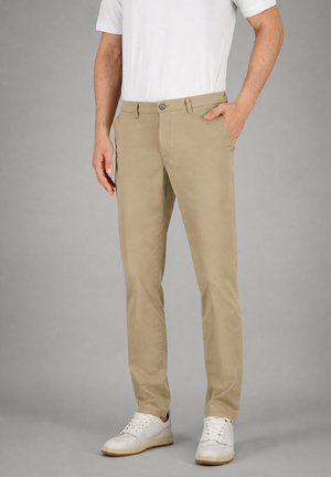 Mann trägt beige Chinos, weiße Sneakers und ein weißes Kurzarmhemd, mit einer Hand in der Tasche vor grauem Hintergrund.