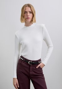 Wit geribd top met lange mouwen en een hoge hals, gecombineerd met bordeauxrode broek met een zwarte riem voorzien van zilverkleurige studs.