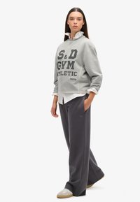 Grijze trui met de opdruk "S&D GYM ATHLETIC", gelaagd over een witte shirt, gecombineerd met donkergrijze broek met wijde pijpen en beige sneakers.