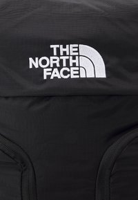 Svart stoff ryggsekk med en fremtredende hvit brodert logo av "The North Face" med et buet design og gitterlignende tekstur.